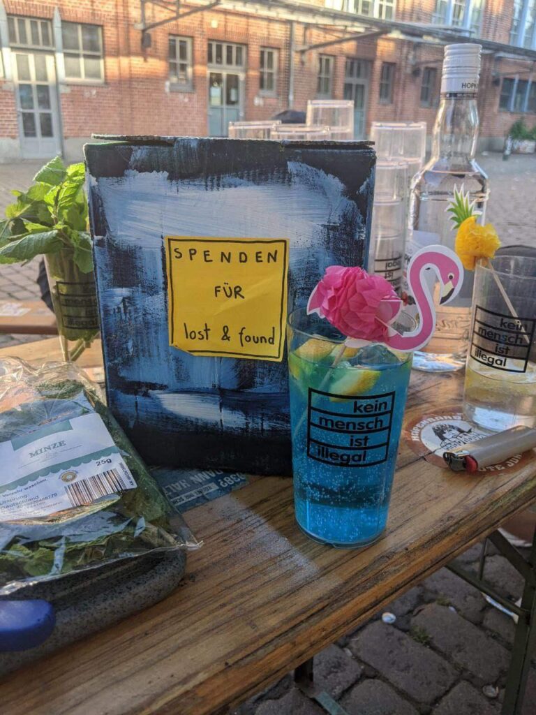 Eine Bierbank vor dem Centro. darauf eine Spendendose in blau und ein Cocktail mit ebenso blauer Farbe. Als Deko ein rosa Flamingo. Daneben noch eine Packung Minze. Es ist noch Tag. 